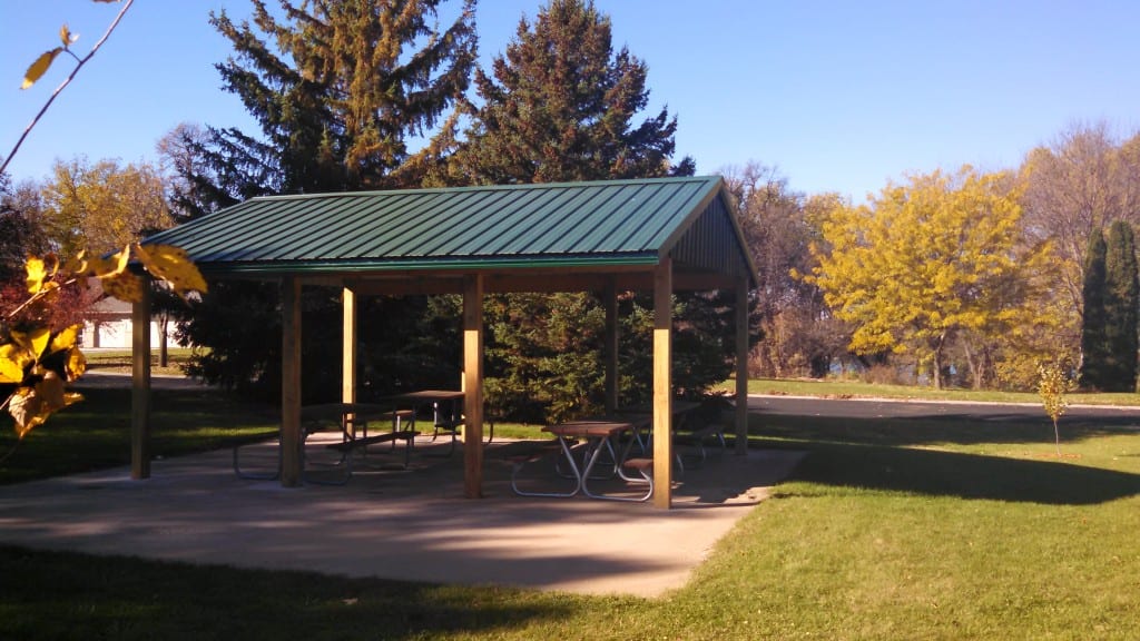 A park shelter with a green metal roof and several picnic tables beneath it sits on a concrete pad, surrounded by grass, trees, and a clear blue sky.