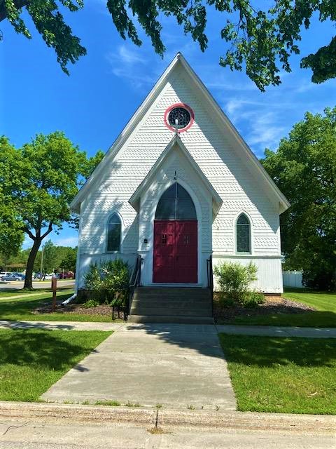 A small white church with a steep roof, red front doors, arched windows, and a circular window above the entrance, surrounded by green grass and trees on a sunny day.