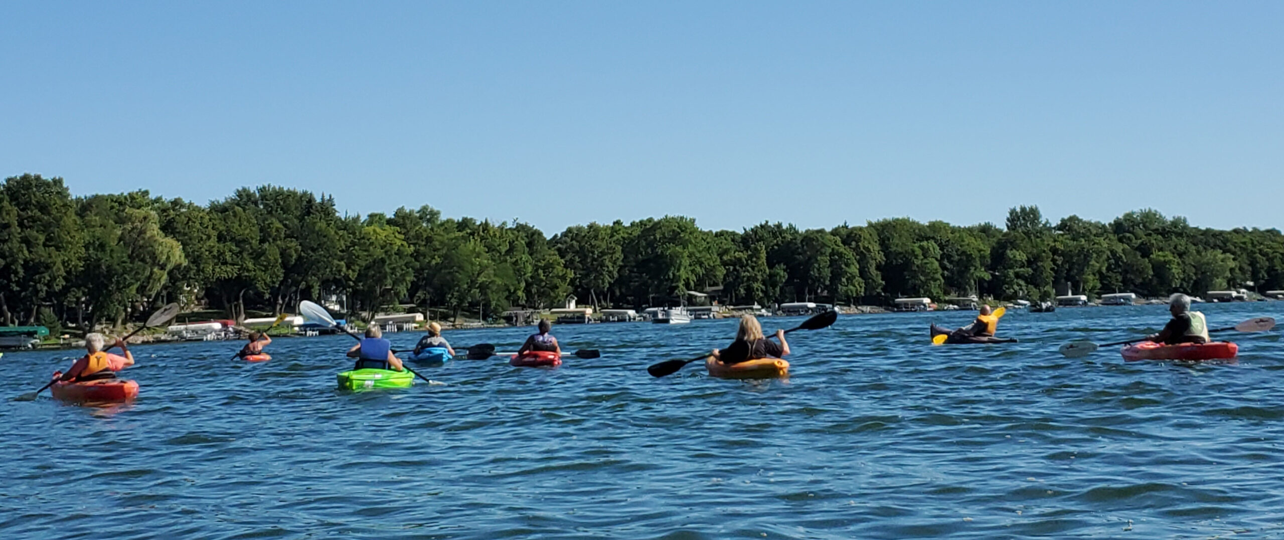 A group of people kayak on a lake under a clear blue sky, with green trees and lakeside houses visible along the shoreline in the background.