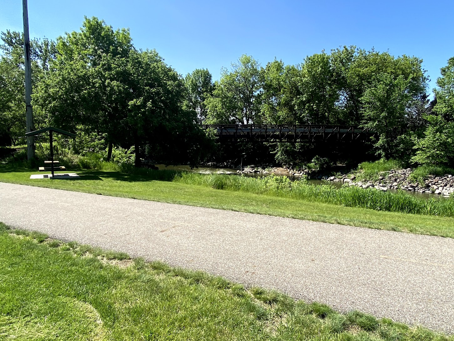 A paved path runs through a grassy park with trees, a small river, and a wooden pedestrian bridge in the background under a clear blue sky.
