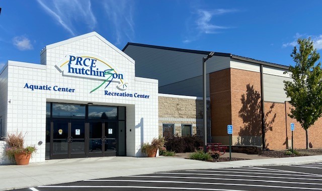 Front entrance of the Hutchinson PRCE Aquatic Center and Recreation Center, with clear signage, large windows, potted plants, accessible parking spots, and a partly cloudy blue sky above.