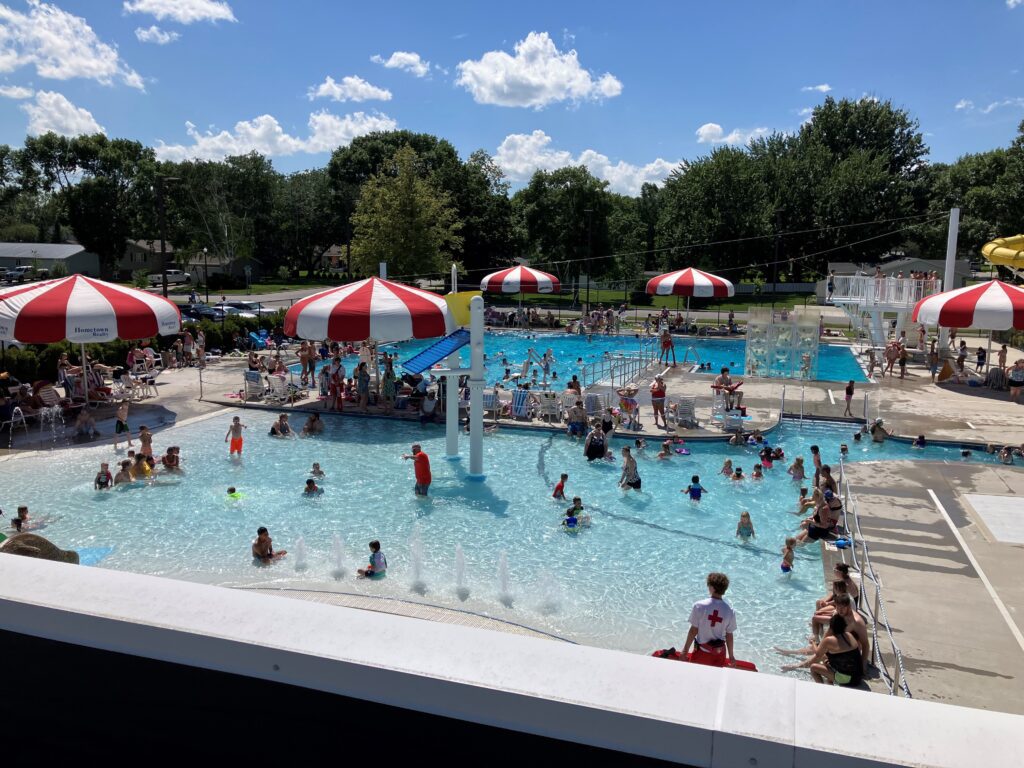 A busy outdoor public pool with people swimming and playing in the water. Red-and-white umbrellas shade seated people. Lifeguards watch from poolside. Trees and blue sky with scattered clouds are in the background.