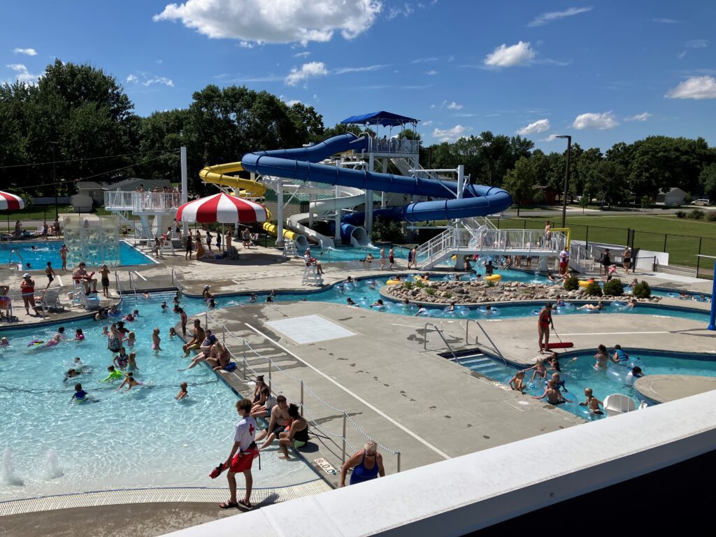 A busy outdoor water park with many people swimming and playing in pools. There are large blue and yellow water slides, a splash pad area, and lifeguards watching over the guests. The weather is sunny with a clear sky.