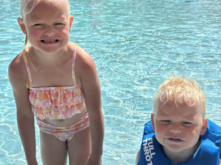 Two young children with light hair smile by the edge of a clear, shallow swimming pool. The girl wears a floral swimsuit, and the boy wears a blue life jacket. Sunlight sparkles on the water around them.